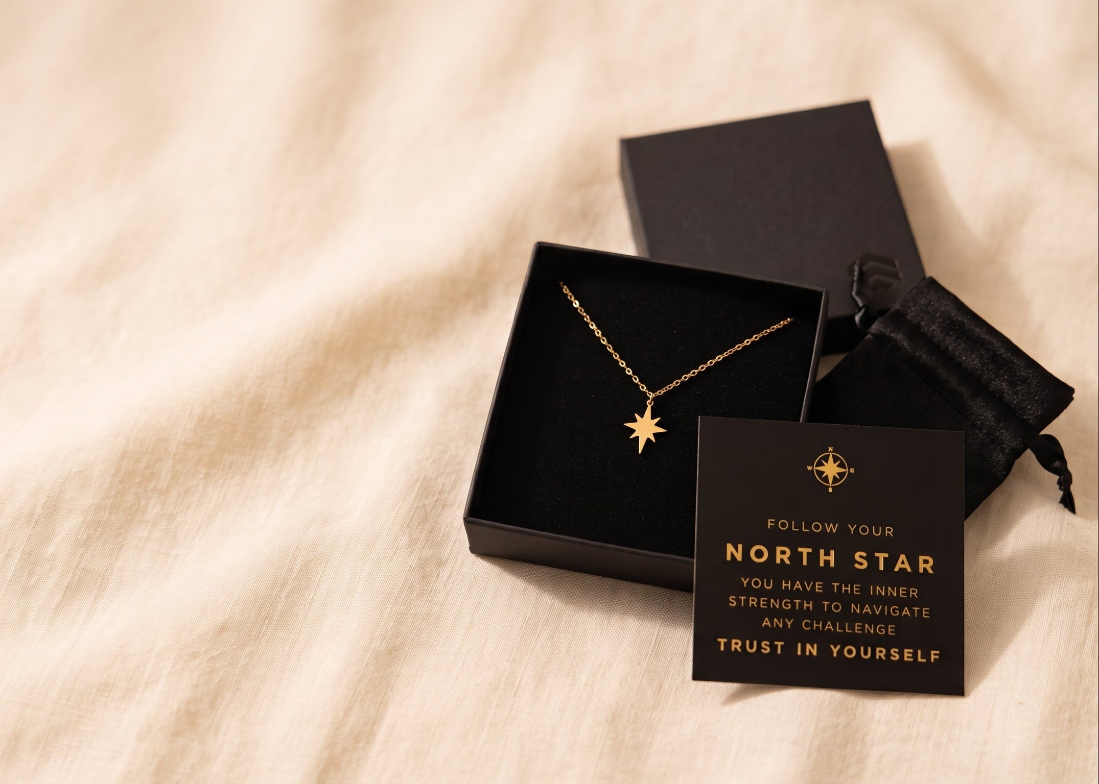 A gold necklace with a compass needle pendant displayed in an open black gift box next to a black card that reads 'NORTH STAR' and 'TRUST IN YOURSELF', set on a beige fabric background.